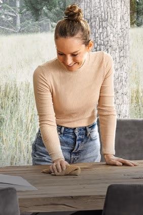 Eine Frau steht über einem Holz Esstisch und hat einen Putzlappen in der Hand.