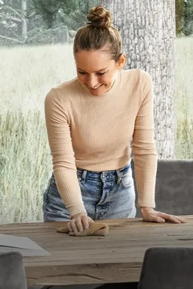 Eine Frau steht über einem Holz Esstisch und hat einen Putzlappen in der Hand.