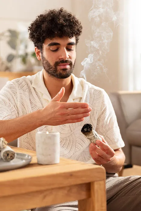 Man sitzt mit einem qualmenden Kräuterbündel in der Hand und räuchert den Raum.