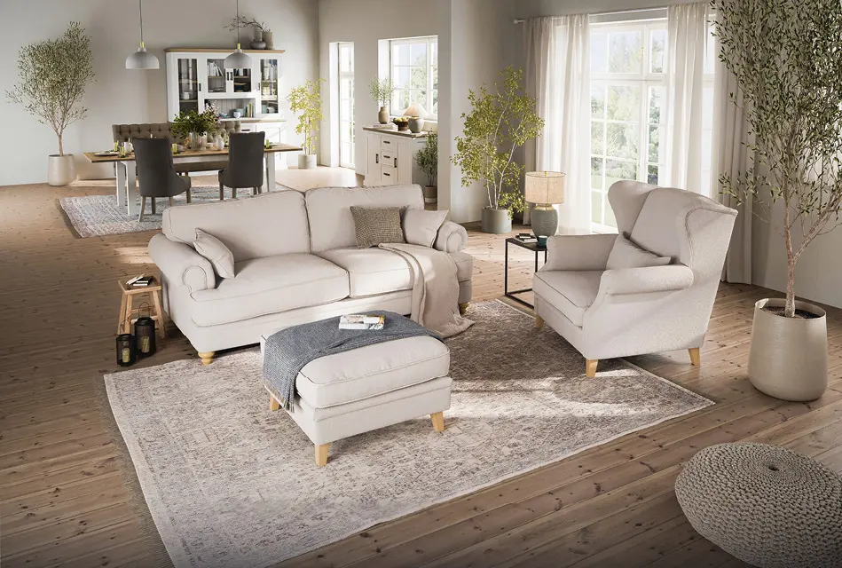 Großzügiges Wohnzimmer mit hellem Sofa, passendem Sessel und Hocker auf Holzfußboden. Offener Übergang zum Essbereich mit Holztisch und Stühlen, große Fenster, natürliche Materialien und wohnliche Einrichtung.