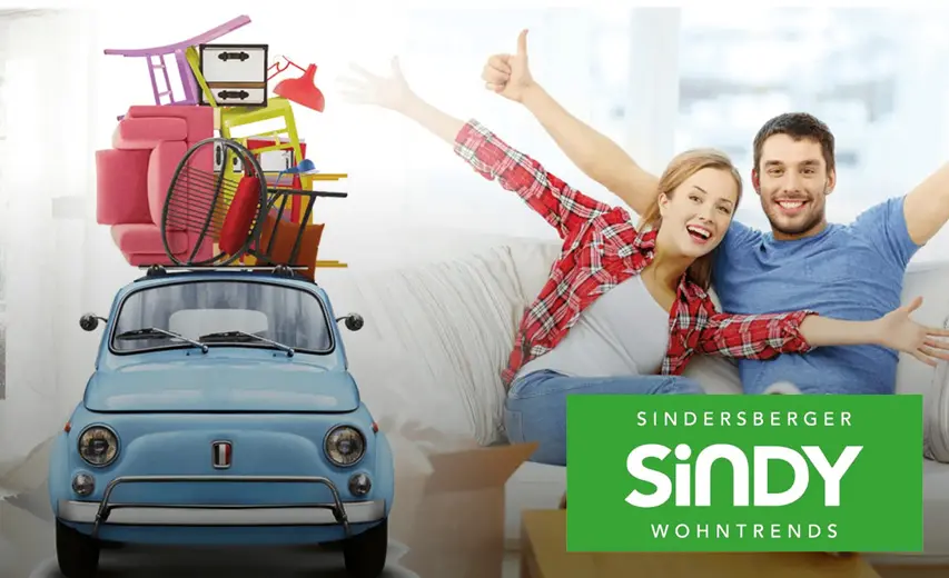 Junges Paar sitzt freudig auf einem Sofa neben einem kleinen Auto voller Möbel – Sindy Wohntrends
