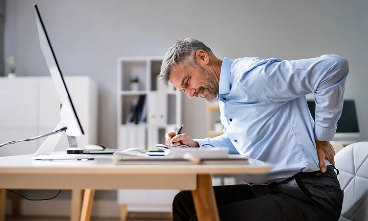 Man am Schreibtisch hebt sich eine Hand an den Rücken und hat einen schmerzlichen Gesichtsausdruck.