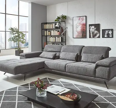 Graues Ecksofa mit Ottoman links, davor schwarzer Couchtisch mit Vase und Schale, links großes Fenster, dahinter Bücherregal und Wandbilder im Wohnzimmer.