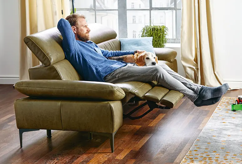 Frau auf naturfarbenem Sofa, Mann und Frau im Wohnbereich mit Holzfußboden, Sessel und komfortabler Couch, Mann mit Hund auf grünem Relaxsofa im Wohnzimmer mit Teppich und Holzfußboden