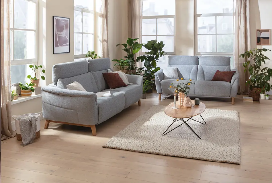 Helles Wohnzimmer mit grauer Sofagruppe, zwei Polstersofas, Couchtisch und Teppich. Große Fenster, Pflanzen und natürliche Materialien schaffen eine wohnliche Atmosphäre mit modernem Einrichtungsstil.