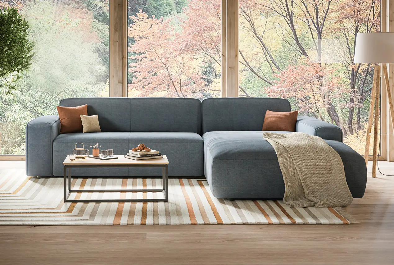 Moderne Wohnlandschaft in Grau mit Recamiere und Kissen, platziert vor bodentiefen Fenstern mit Blick ins Grüne. Stilvolles Sofa im hellen Wohnzimmer mit Teppich und Couchtisch als Inspiration für modernes Wohnen.