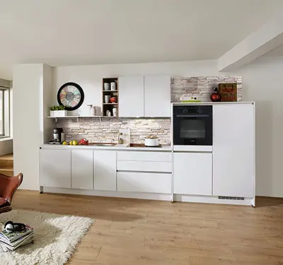Gerade Einbauküche in Weiß mit Hochschrank und Backofen rechts, davor Holzarbeitsplatte, helle Maueroptik Nischenrückwand, links Uhr und offene Regale.