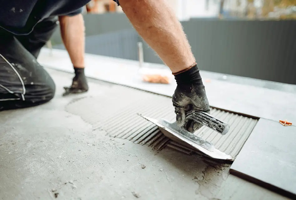 Ein Handwerker trägt mit einer Zahnkelle Fliesenkleber auf den Boden auf. Die Szene zeigt professionelle Bodenverlegung, Untergrundvorbereitung und präzise Handwerksarbeit im Innenausbau.