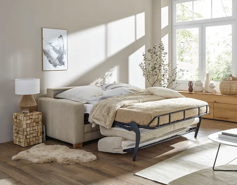 Ausgeklappte Schlafcouch in Naturtönen mit Nachttisch, auf Holzboden im hellen Raum