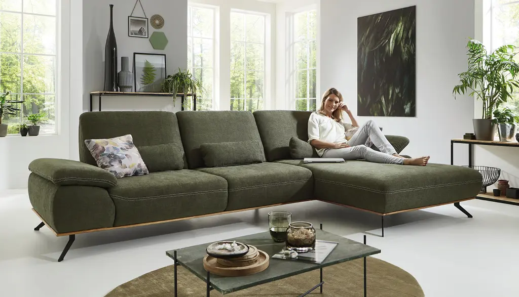 Großes Ecksofa in Olivgrün von Couchliebe mit filigranen Metallfüßen in hellem Wohnzimmer, auf dem eine Frau entspannt liest – erhältlich bei Möbel Rommel.
