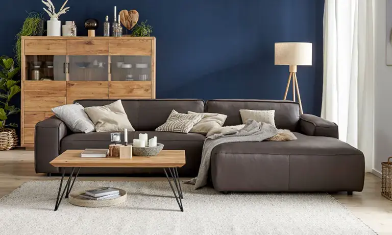 Dunkelbraunes Ecksofa vor dunkelblauer Wand, mit Holz Couchtisch im Vordergrund und Sideboard links auf hellem Teppich