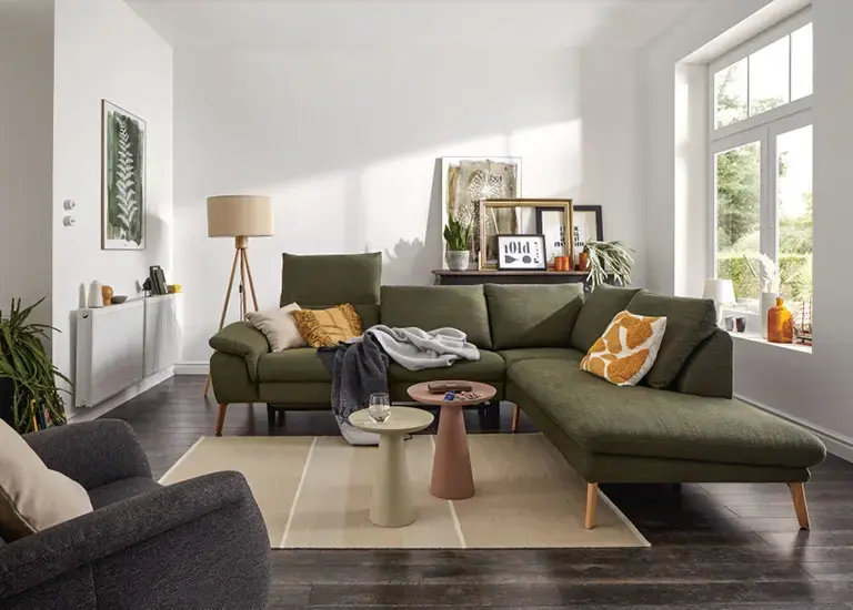 Wohnzimmer mit grünem Ecksofa, Kissen und Decke, davor zwei runde Beistelltische auf hellem Teppich, rechts grosse Fensterfront mit Blick ins Grüne.