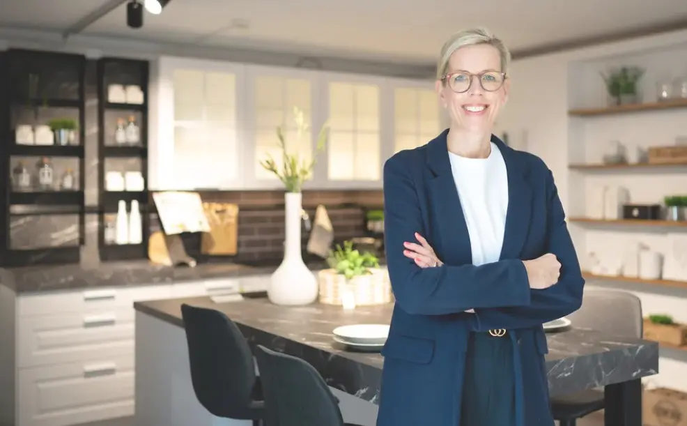 Frau mit Brille und verschränkten Armen steht vor Kücheninsel mit Stühlen, im Hintergrund moderne Einbauküche mit Regalen und Beleuchtung