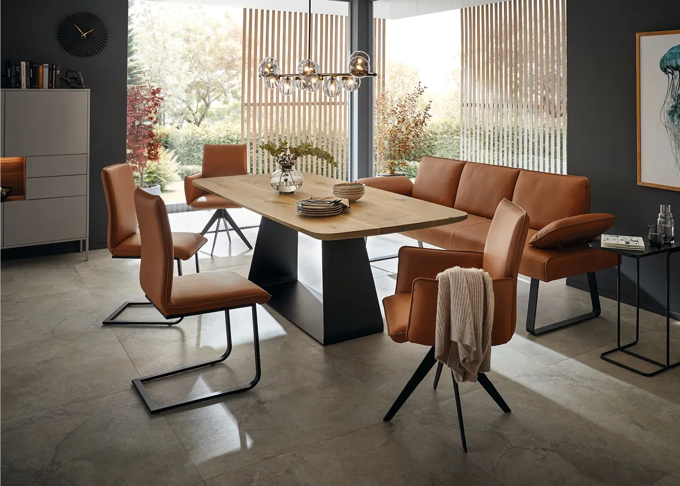 Lesender Mann auf braunem Stuhl an Esstisch aus Holz vor einem großem Fenster in hellem Esszimmer