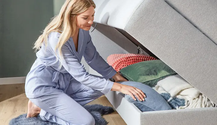 Frau kniet vor geöffnetem Bettkasten eines grauen Boxspringbetts, elektrisch verstellbar