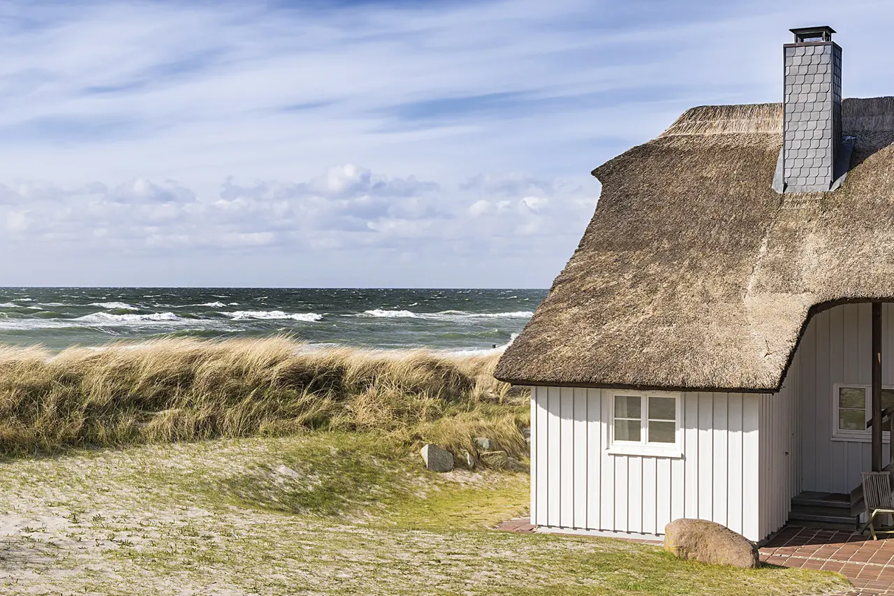Kleines Reetdachhaus in heller Holzbauweise steht direkt hinter den Dünen an der Nordsee mit Blick auf Meer, Strandgras und Wolkenhimmel und vermittelt Küstennähe, Ruhe und naturnahes Wohnen