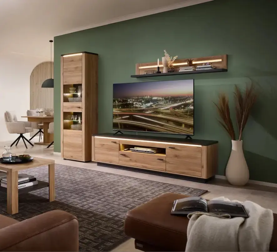 Wohnzimmerausstellung mit grüner Wand, Holz Wohnwand und großem Fernseher, links Essbereich mit Tisch und Stühlen, vorn Couchtisch und Ledersitz.