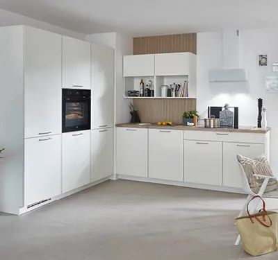 L förmige weiße Einbauküche mit hoher Schrankwand links und Backofen, rechts Unterschränke mit Holzarbeitsplatte, offene Regale und Wandhaube vor heller Wand.