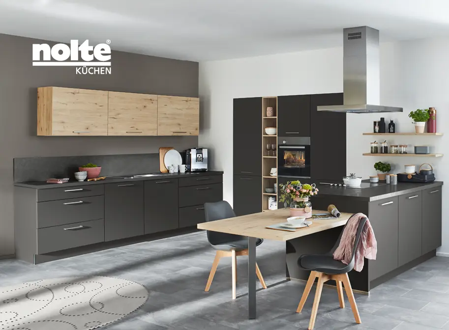 Matt schwarze Küchenzeile mit in der Wand eingelassenem Schrank mit integriertem Backofen. An dem Küchenblock ist eine integrierte Sitzgelegenheit mit zwei Stühlen.