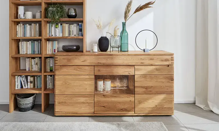 Sideboard aus Eiche mit Glaseinsatz rechts neben hohem Bücherregal, dekoriert mit Vasen und Kerzen vor heller Wand im Wohnzimmer