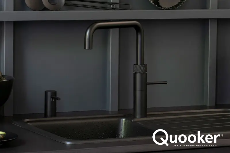 Schwarzer Quooker Wasserhahn ueber Spuelbecken in dunkler hoelzerner Designkueche und Quooker Logo