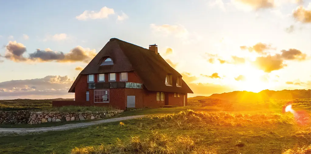 Traditionelles Reetdachhaus in ruhiger Küstenlandschaft mit Wiesen und Dünen, warmes Abendlicht bei Sonnenuntergang vermittelt Erholung, Naturverbundenheit und Urlaub an der Nordsee