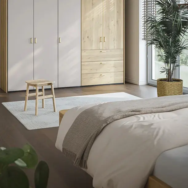 Schlafzimmer mit Bett im Vordergrund, Kleiderschrank mit Holz und hellen Fronten, Hocker und Pflanze am Fenster