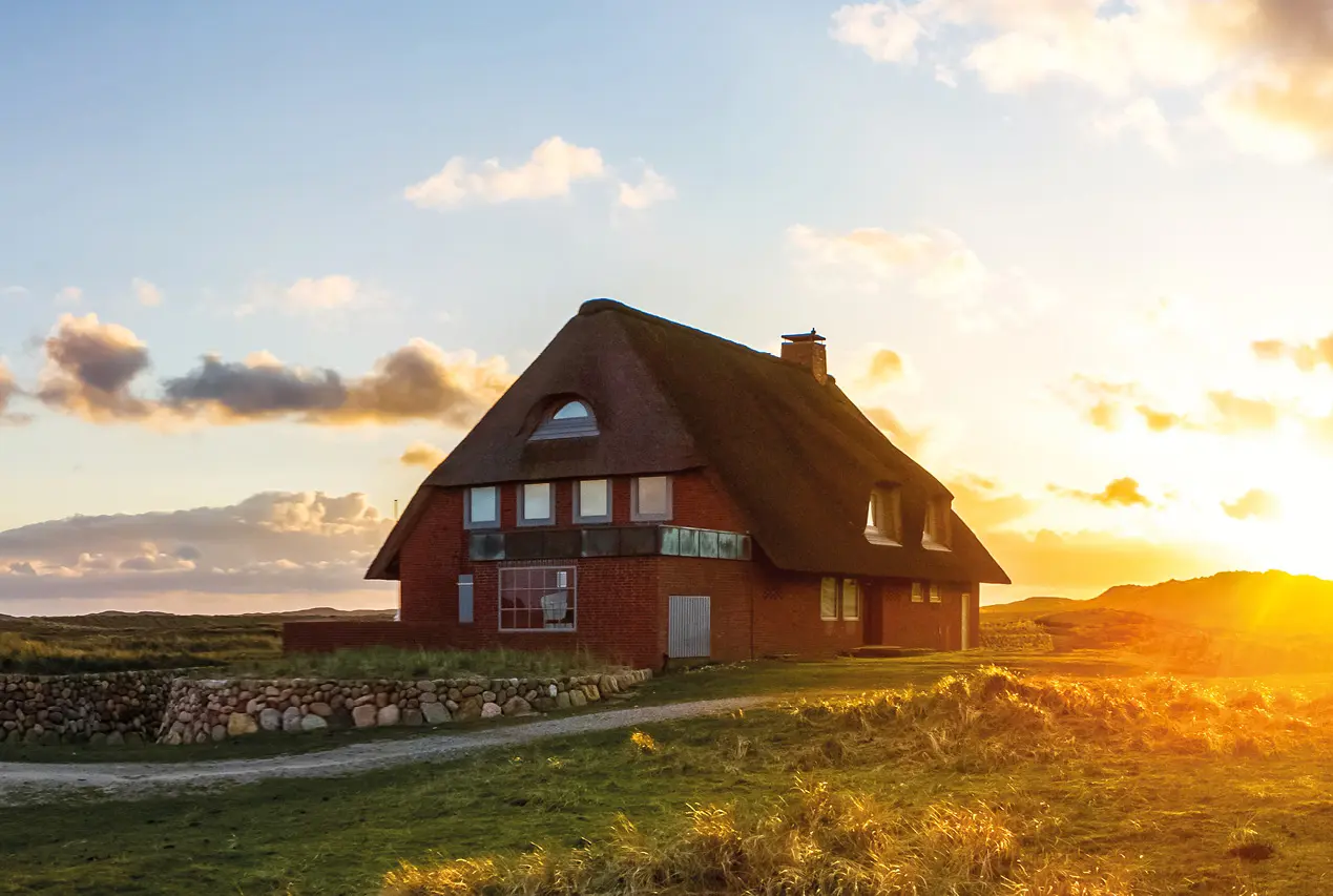 Traditionelles Reetdachhaus in ruhiger Küstenlandschaft mit Wiesen und Dünen, warmes Abendlicht bei Sonnenuntergang vermittelt Erholung, Naturverbundenheit und Urlaub an der Nordsee
