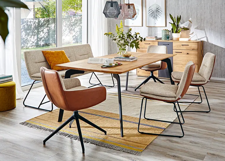 Abverkauf: Männer tragen Sessel im Möbelhaus, Holztisch mit Stühlen in Naturfarben, Mann auf sandfarbenem Sessel sitzend