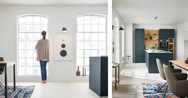 Großraum Wohnung mit hohen Fenstern, einer Frau die nach draußen blickt. Eine weite Küche rechts.