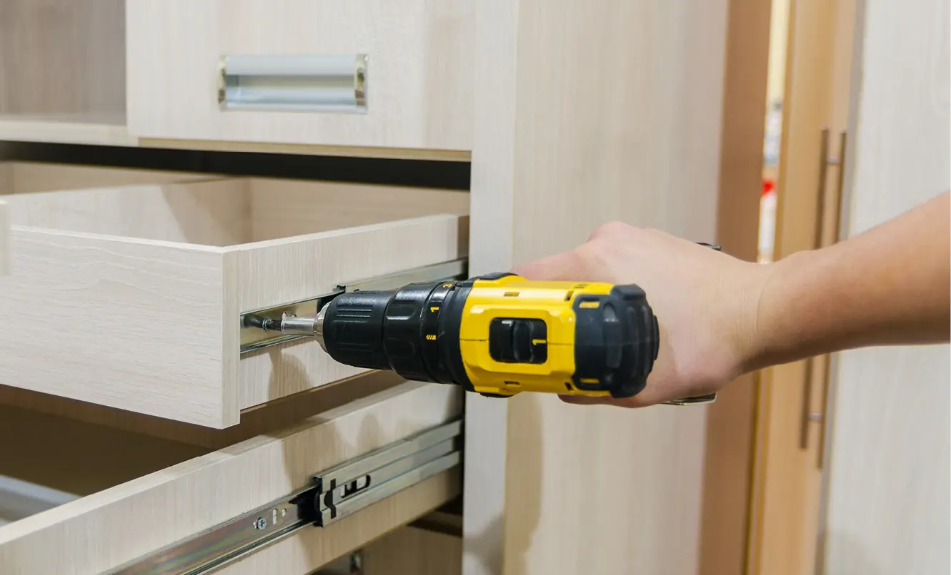 Hand mit Akkuschrauber befestigt die Schiene eines geöffneten Schubkastens an einem Schrank aus hellem Holz.