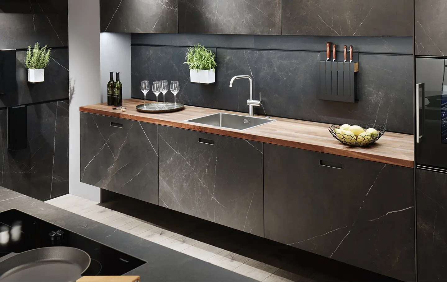 Elegante Kuechenzeile in dunkler Marmoroptik mit Holzarbeitsplatte und an Wand befestigter Messerblock und Krauetertopf.