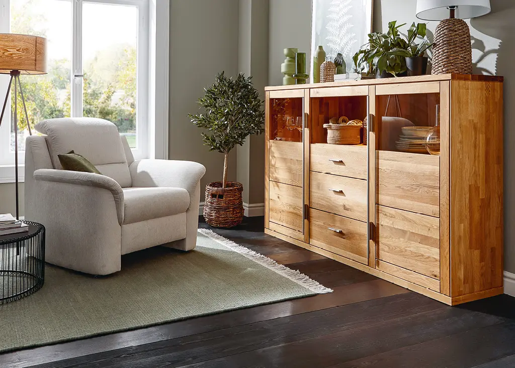 Hölzernes Highboard mit Schubladen und Vitrine neben grauem Sessel im Wohnzimmer
