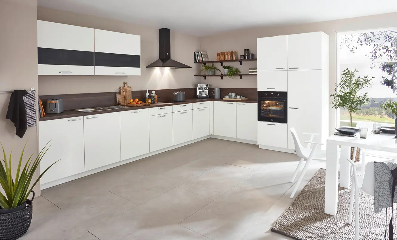 Weisse L förmige Einbauküche mit dunkler Arbeitsplatte, Wandregalen und Hochschrank mit Backofen, rechts weisser Esstisch mit Stühlen vor grossem Fenster und Pflanze.