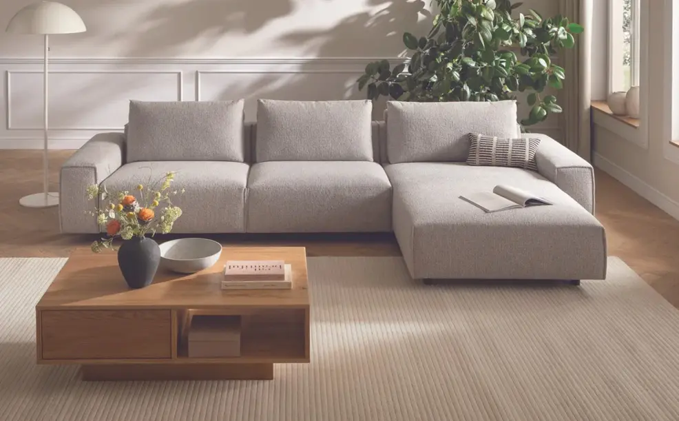 Helles Ecksofa mit Kissen, Couchtisch aus Holz davor, Teppich und Pflanze neben Fenster im Wohnzimmer