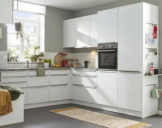  Weiße Eckküche mit Backofen, hohen Schränken und grauer Arbeitsplatte, vor Fenster mit Pflanzen und hellem Teppich.