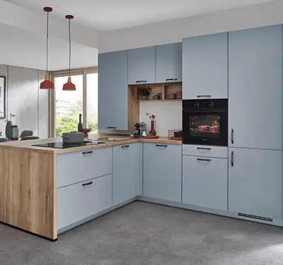 L förmige hellblaue Einbauküche mit Holzarbeitsplatte und niedriger Theke links, Hochschrank mit Backofen rechts, grauer Boden, zwei rote Pendelleuchten vor Fenster.