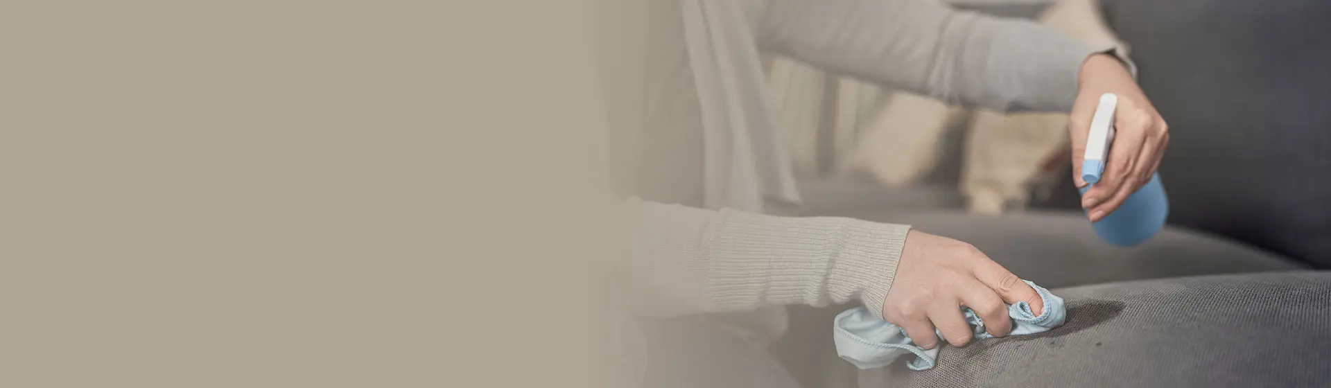 Hand sprüht Reinigungsmittel auf graues Polster und wischt mit Tuch über die Oberfläche eines Sofas