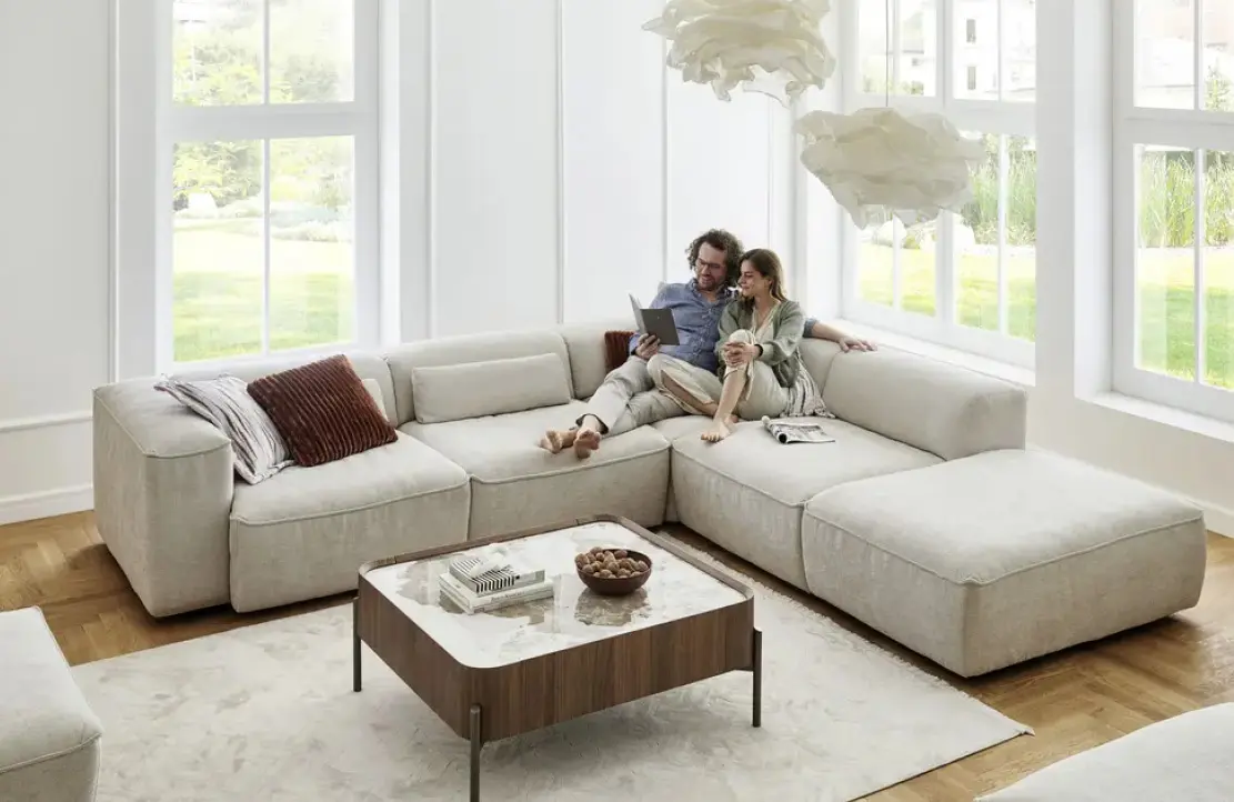Helles Ecksofa vor hoher Fensterfront, zwei Personen sitzen in der Mitte, davor Couchtisch mit Holzrahmen und heller Platte auf Teppichboden über Parkett.