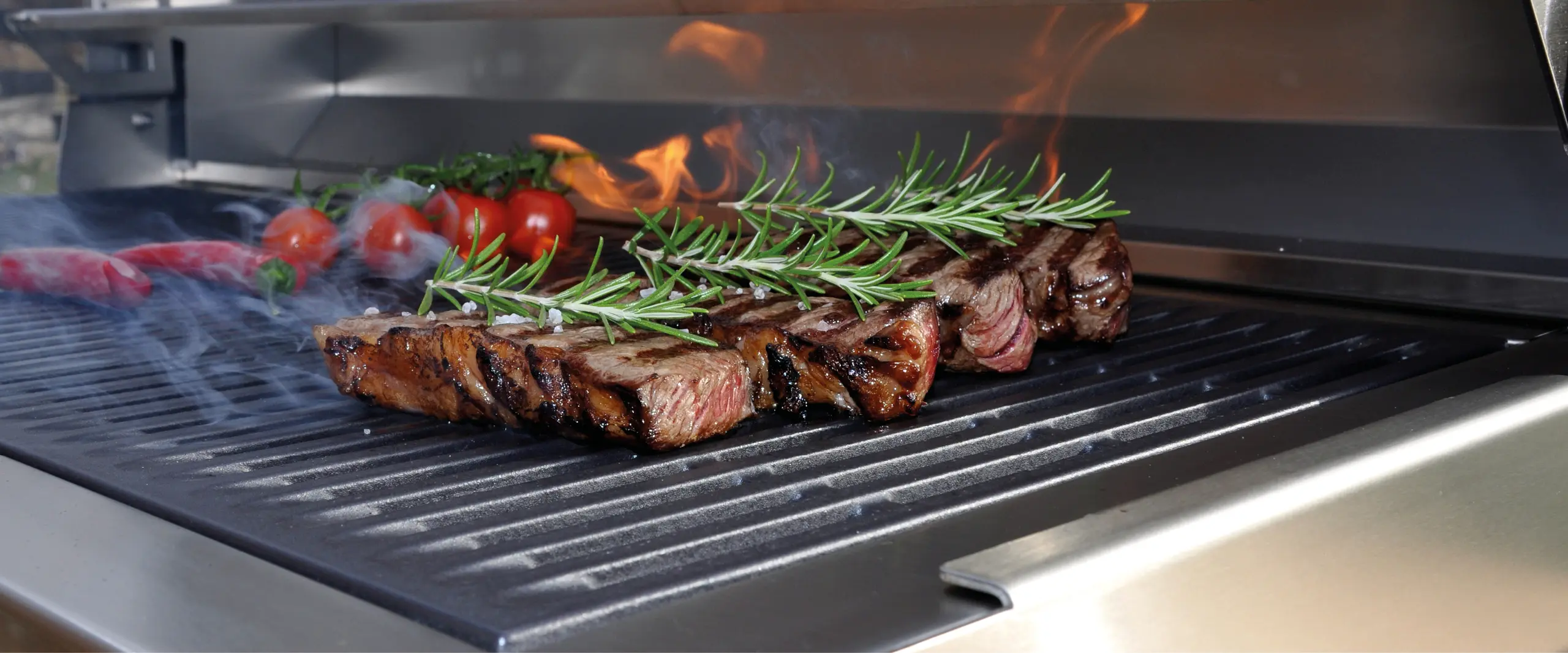 Steaks und Kirschtomaten auf Grillflaeche einer Flammkraft Outdoor-Kueche