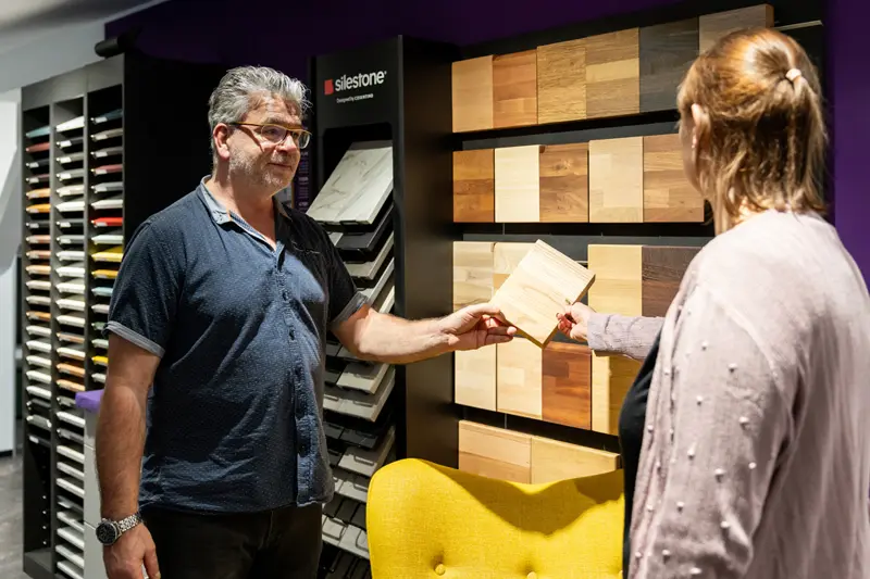 Beratungsgespraech mit Kundin vor Regal mit Materialmustern fuer die Kuechenplanung.