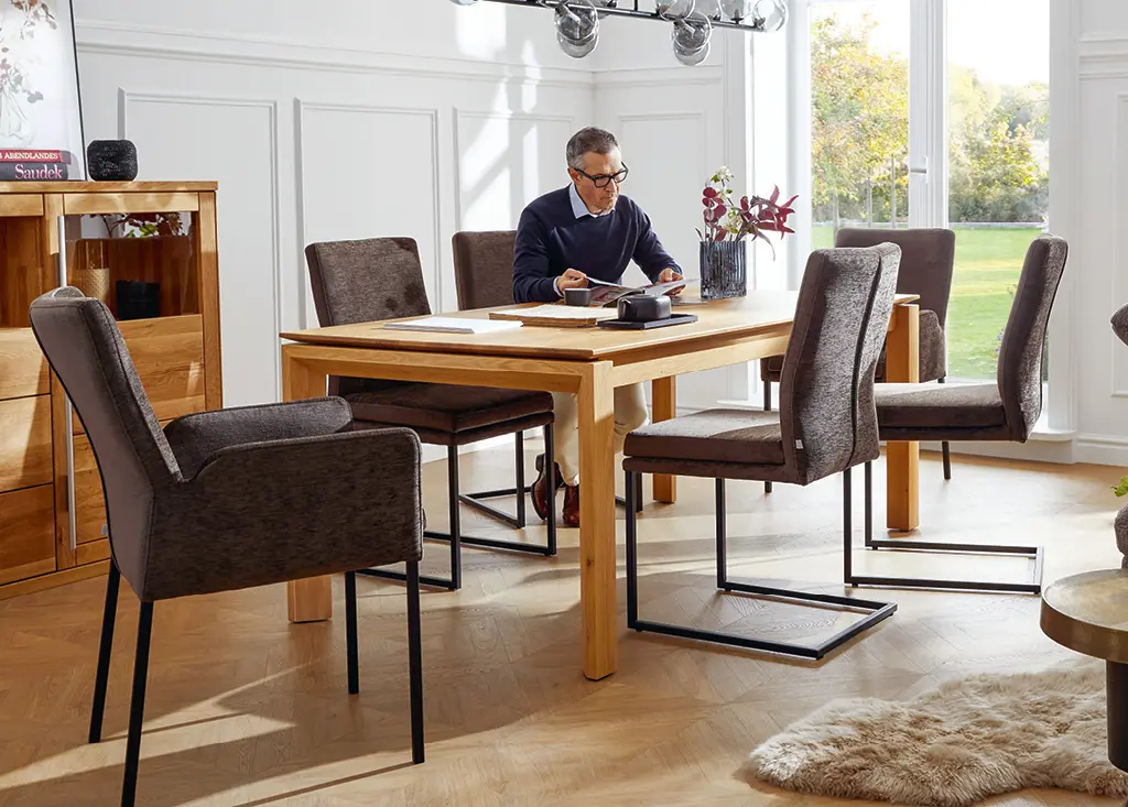 Lesender Mann auf braunem Stuhl an Esstisch aus Holz vor einem großem Fenster in hellem Esszimmer