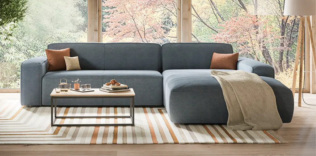 Moderne Wohnlandschaft in Grau mit Recamiere und Kissen, platziert vor bodentiefen Fenstern mit Blick ins Grüne. Stilvolles Sofa im hellen Wohnzimmer mit Teppich und Couchtisch als Inspiration für modernes Wohnen.