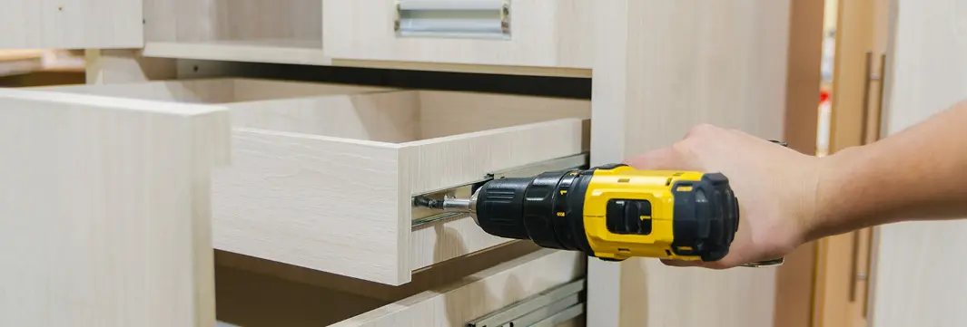 Hand mit Akkuschrauber befestigt die Schiene eines geöffneten Schubkastens an einem Schrank aus hellem Holz.