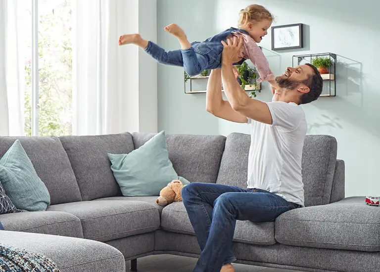 Vater hebt lachendes Kind in die Luft vor grauem Sofa im hellen Wohnzimmer. Familienmoment mit moderner Wohnzimmereinrichtung und gemütlicher Wohnlandschaft
