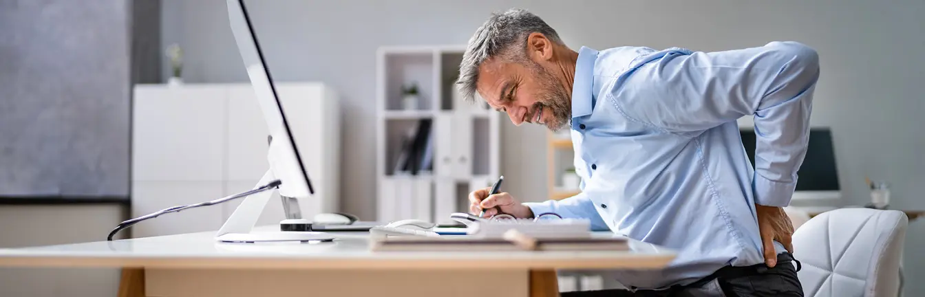 Man am Schreibtisch hebt sich eine Hand an den Rücken und hat einen schmerzlichen Gesichtsausdruck.