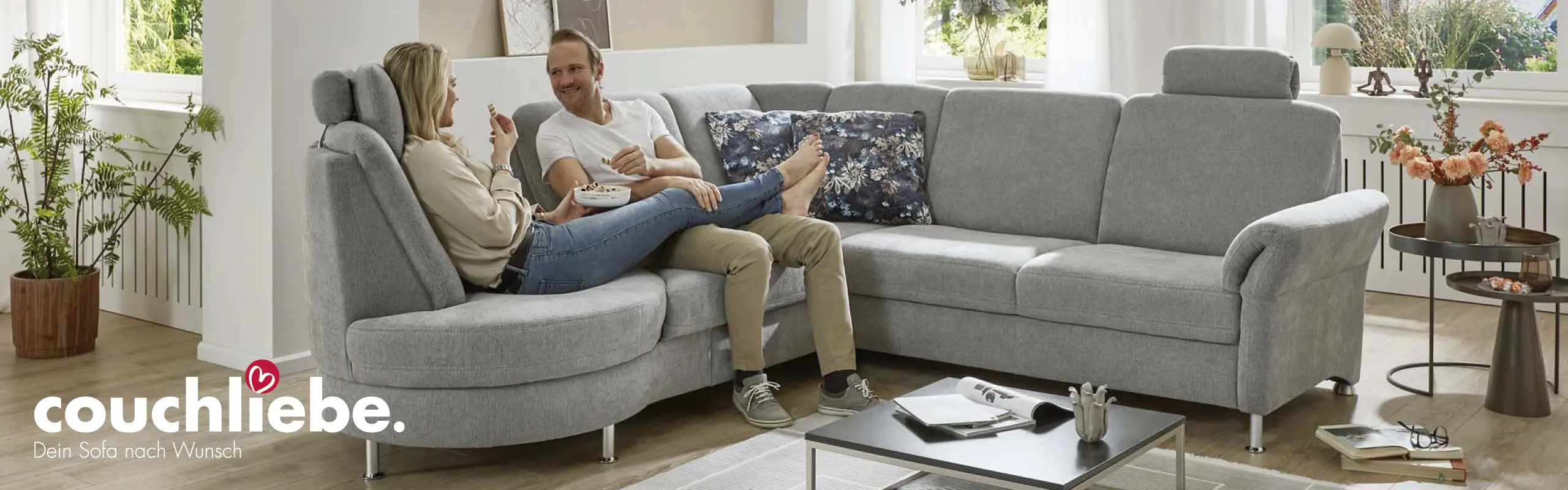 Modernes graues Ecksofa von Couchliebe in hell eingerichtetem Wohnzimmer, auf dem ein Paar entspannt sitzt – erhältlich bei Möbel Rommel.