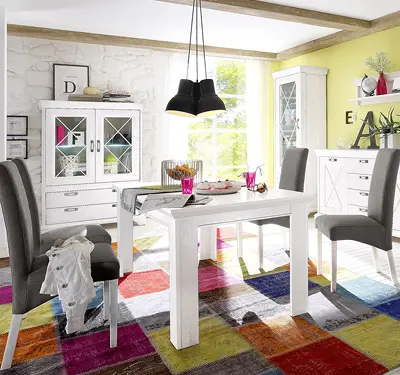 Helles Esszimmer mit weißem Esstisch, vier grauen Stühlen und zwei weißen Vitrinen, darüber schwarze Pendelleuchten, bunter Patchwork Teppich auf dem Boden.