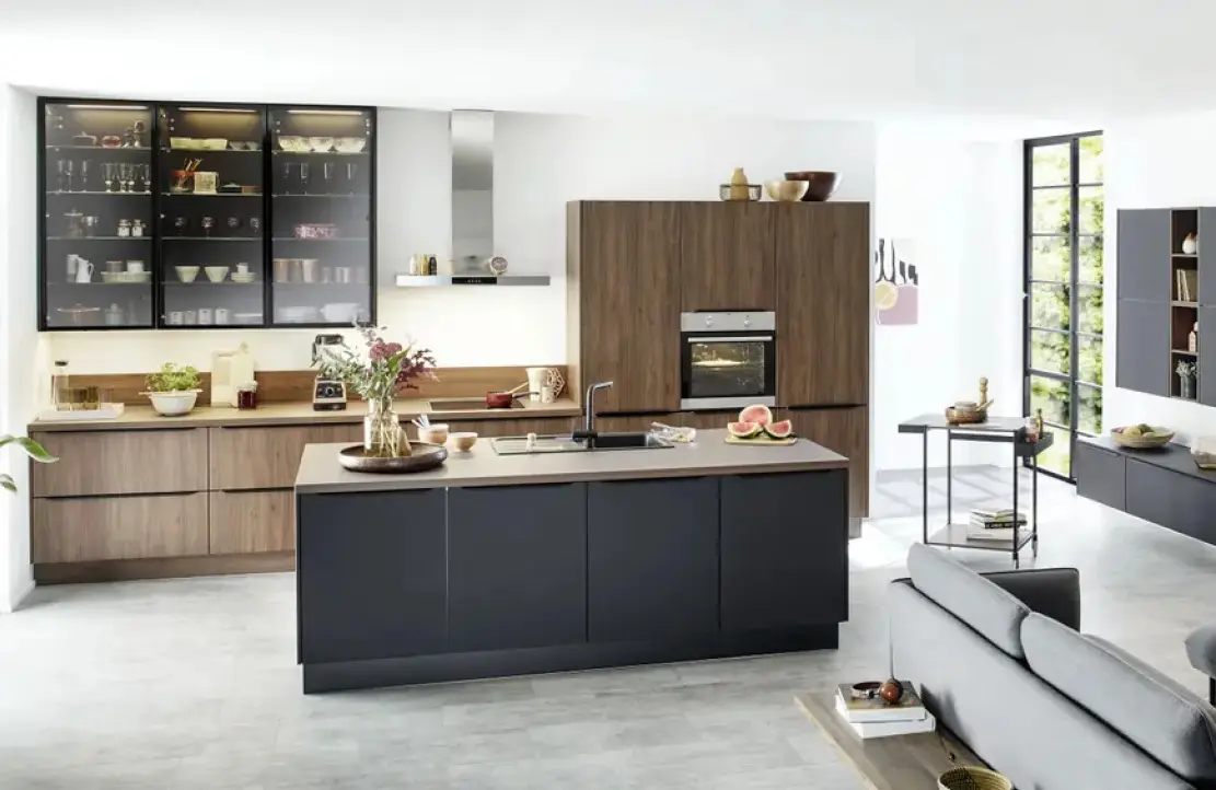 Offene Küche mit dunkler Insel, Holzfronten und hohem Schrank mit Backofen, links Glasvitrinen, dahinter Arbeitszeile, rechts Fensterfront und Sideboards.