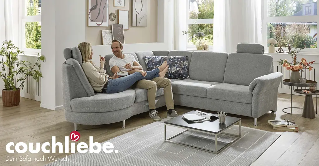 Modernes graues Ecksofa von Couchliebe in hell eingerichtetem Wohnzimmer, auf dem ein Paar entspannt sitzt – erhältlich bei Möbel Rommel.